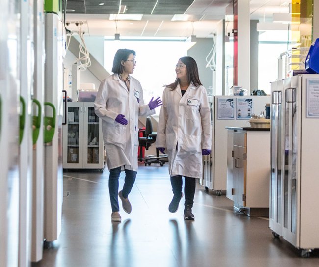 Two lab technicians in white lab coats having a conversation while walking in lab