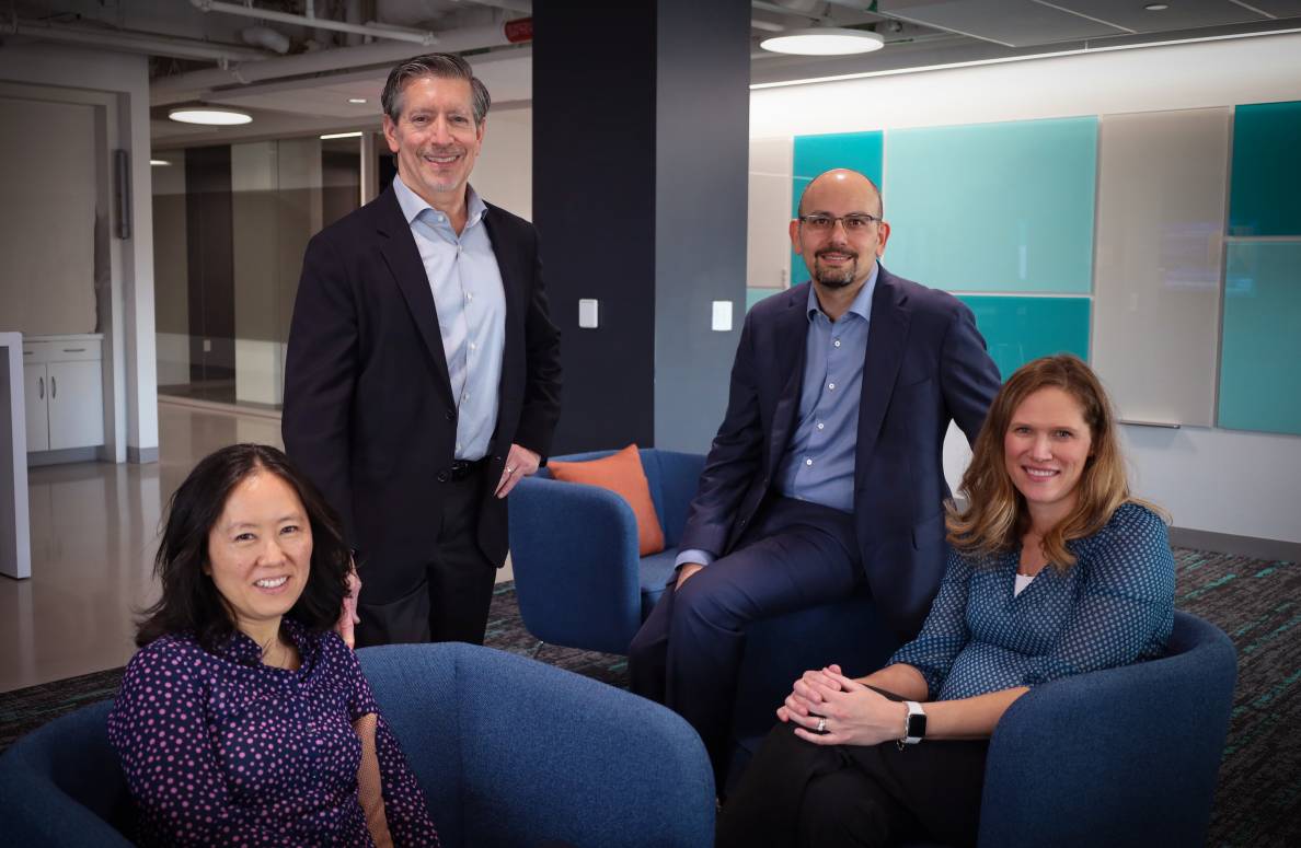 Four people in a modern office setting, with two seated on blue chairs and two standing behind them