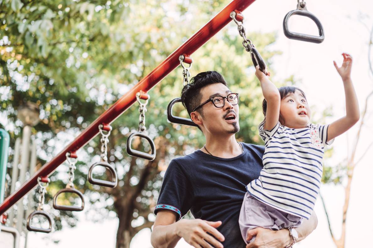 Adult holding a child while she hands on monkey bar