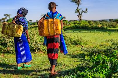 two individuals in field carrying large yellow container on their back.