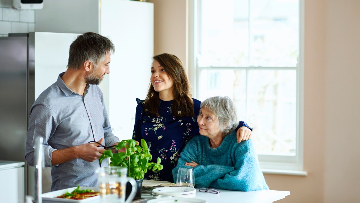 younger woman in the kitchen with her arm around an older woman and a young man nearby smiling