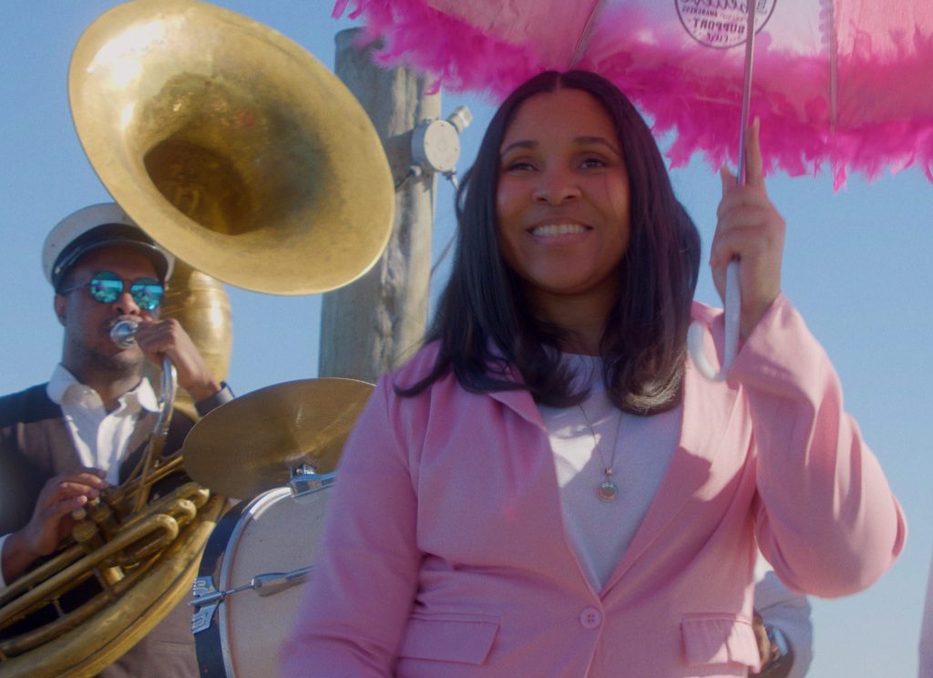 woman smiling and walking with a pink umbrella and man behind her playing a tuba