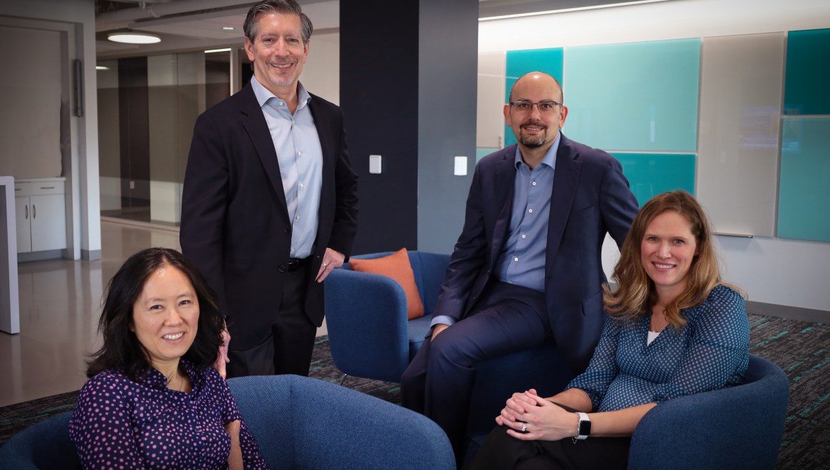 Four people in a modern office setting, with two seated on blue chairs and two standing behind them